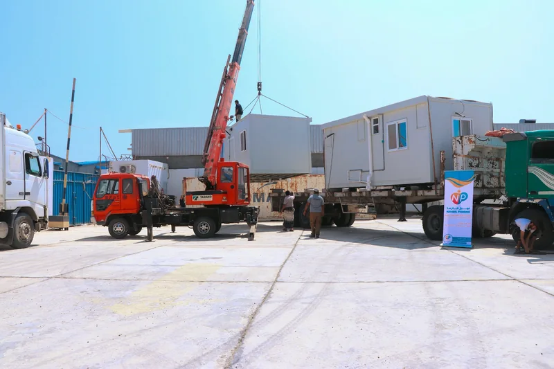 A red crane lifting a prefabricated unit beside a truck carrying mobile clinic units