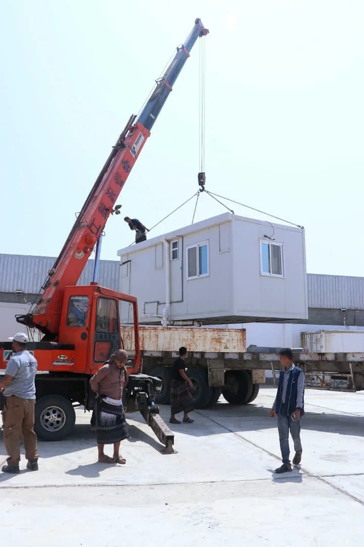 A clinic unit suspended by crane above a transport trailer with people on site