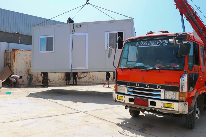 A grey unit with a door and two windows suspended from lifting slings behind a red crane truck